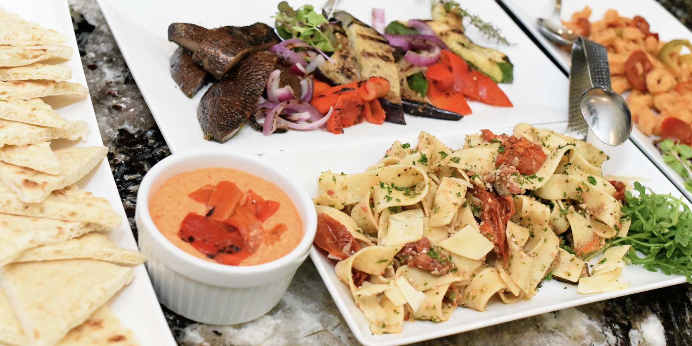 Catering spread with pasta, grilled vegetables, pita bread, and roasted red pepper dip